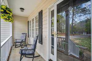 A cozy front porch featuring two patterned rocking chairs, a black door with a glass pane, and a hanging basket with yellow flowers. The porch overlooks a yard with trees lining the background, creating a peaceful, welcoming atmosphere.