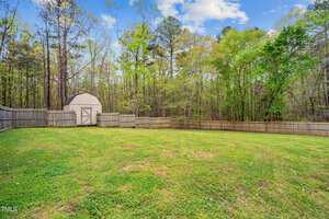 A spacious, grassy backyard with a wooden fence surrounding the area. A small, white garden shed is in the corner. Tall trees border the yard, and the sky is partly cloudy.