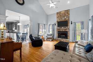 A spacious living room with wooden floors, featuring a stone fireplace, large windows, and neutral-colored seating. A ceiling fan hangs overhead, while an adjacent open kitchen with gray cabinets is visible. A piano sits to the left.