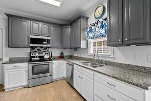 A modern kitchen with gray upper cabinets and white lower cabinets, granite countertops, and stainless steel appliances. A blue and white floral valance hangs over the window. The backsplash is black, white, and gray tile. A round wall clock is above.
