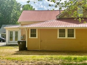 A yellow house with a red metal roof and white-framed windows. A black trash bin is outside next to the wall. The yard is grassy with some bare patches, and a tree casts a shadow on the house. Another small building is visible in the background.