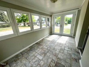A sunlit room with large windows and a glass door, featuring stone tile flooring. The windows reveal a view of a residential street with trees and parked cars outside. The room has light-colored walls and a ceiling light fixture.