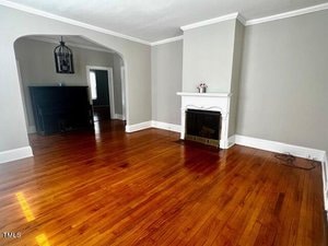 A spacious living room with polished hardwood floors, light gray walls, and white trim. It features a white fireplace with a mantle. An archway leads to a darker area with a black piano and a pendant light fixture.