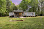 The single-story gray house at 2205 Lauren Mill Drive in Oxford features a charming covered porch, surrounded by a well-maintained lawn and trees. A gravel driveway leads to the carport on the right, all beneath a sky tinged with pink, signaling sunset or sunrise.