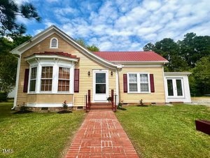 A charming, single-story house on 517 Coggeshall Street features tan siding and a red metal roof. With a prominent bay window, red shutters, and a brick pathway leading to the front door, it sits in a lush green yard framed by Oxford's cloudy blue sky.