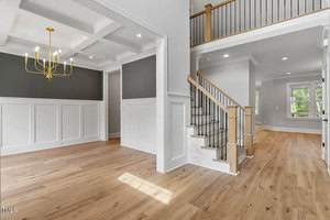 Spacious interior with light wood floors, white paneled walls, and dark gray accents. A staircase with wood and metal railing leads to an upper level with a balcony. A room features a large, modern, gold chandelier. Large windows provide natural light.
