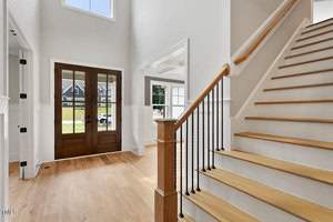 A bright and spacious entryway with wooden flooring, featuring a staircase with wooden handrails and black balusters. Double wooden doors with glass panels lead to the outside, and a large window illuminates the space with natural light.