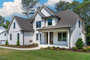 A modern white two-story house with dark roof shingles and a covered front porch. The house has large windows and is surrounded by landscaped greenery with small shrubs and a manicured lawn. A cloudy blue sky is in the background.