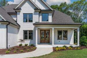 A modern house with a white brick exterior and black trim. It features large wooden front doors, ample windows, and a covered porch. There's a neatly landscaped yard with small shrubs and a concrete walkway leading to the entrance.