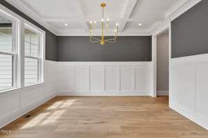 A bright, empty dining room with large windows, dark gray accent walls, white wainscoting, and a light wood floor. It features a modern gold chandelier and a coffered ceiling. Natural light fills the space.