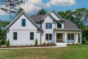 A modern, two-story white house at 991 Browning Place in Youngsville boasts a dark roof, large windows, and a wooden front door. It features a well-maintained lawn and small shrubs lining the walkway. The sky is partly cloudy, with tall trees surrounding the property.