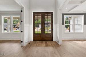 The image shows a bright, empty interior space with light wooden floors and white walls. A large, dark wooden door with glass panes forms the center, allowing a view of a suburban street outside. Flanking windows let in natural light.