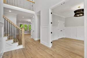 Spacious, modern interior of a home featuring light wood flooring, a staircase with a black handrail, white walls with decorative paneling, and a room with a large, circular black pendant light. Bright windows provide natural light.