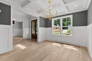 A bright dining room with light wood floors and white wainscoting. A modern gold chandelier hangs from a coffered ceiling. Large windows provide natural light, and a wooden front door is visible in the background.