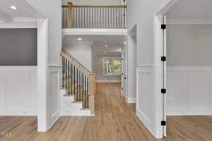 A spacious, modern interior features light wood flooring, white walls, and black railing on a staircase leading to the upper level. The area is well-lit with natural light from windows in the background.