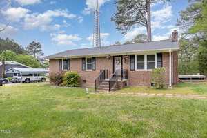 A brick single-story house with a gray roof and white trim set in a grassy yard. Steps lead up to the front entrance. Tall trees surround the property, and a tall radio tower is visible in the background. Blue sky with scattered clouds.