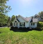 Situated at 3142 Pier Pointe Drive in the charming Creedmoor area, this white single-story house boasts gray shingles and dark shutters. It's surrounded by a lush green lawn and trees, with a large arched window gracing the front. Bushes line the exterior under a clear, sunny sky.