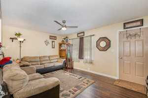 A cozy living room with two beige sofas, a wooden cabinet, and wall decorations. There's a ceiling fan, a window with sheer curtains, and a light-colored door. A patterned rug is on the dark hardwood floor.