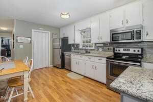 A modern kitchen with white cabinets, granite countertops, and stainless steel appliances. There's a dining table with chairs on a wooden floor, and a window above the sink provides natural light.
