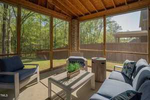 A cozy outdoor screened porch with comfortable blue cushioned seating set around a white coffee table. A potted plant sits on the table. The porch overlooks a green yard through large screened windows, framed by wooden beams.