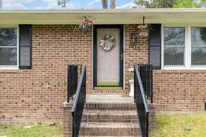 A brick house with a black door and decorative wreath. Two black railings lead up the steps to the entrance. Windows with black shutters flank the door, and potted flowers hang beside it.