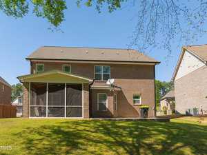Back view of a two-story brick house with a screened porch. The yard has green grass, and trees are visible in the background. A satellite dish is mounted on the house.