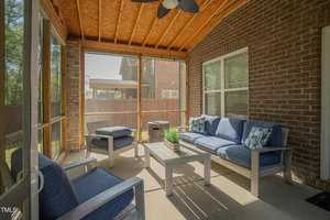 A cozy screened-in porch features a wooden ceiling and brick walls. It is furnished with blue cushioned chairs, a sofa, and a wooden coffee table with a small potted plant. Sunlight filters through the screens, casting shadows on the floor.