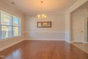 A bright, empty room with hardwood floors, a chandelier, and a large window. There's a rectangular mirror on the wall and a doorway leading to another area. Natural light fills the space, highlighting the neutral wall colors.