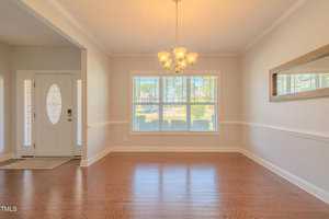 A bright, empty room features wooden floors and a large window with white blinds, letting in natural light. A chandelier hangs from the ceiling. There's a white front door to the left and a mirror on the right wall.
