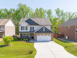 Nestled on 1496 Fireside Lane, this two-story Creedmoor house boasts a gray and white exterior, a double garage, and a wide driveway. It's surrounded by lush green grass and small shrubs, with neighboring brick homes and a line of trees providing a picturesque backdrop.