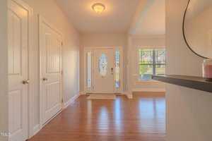 A bright entrance hallway with wooden floors, white walls, a central light fixture, and a glass-paneled white front door. There are two closed doors on the left and a rectangular mirror on the right wall.