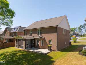 A two-story brick house with a screened porch in a suburban neighborhood. The backyard features a grassy lawn and a concrete patio. Adjacent houses and a clear blue sky are visible in the background.