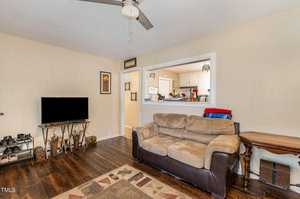 A cozy living room with a beige sofa, a flat-screen TV on a stand, and a ceiling fan. Wooden floor with a patterned rug. Open view into a kitchen with white cabinets. Wall art and a shoe rack are visible.