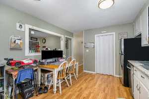 A kitchen and dining area with wooden floors, white cabinets, and stainless steel appliances. A dining table is set with two computers and surrounded by chairs. Shelves hold various items, including a toy car. Walls are decorated with pictures and plants.