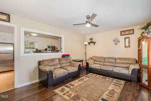 A cozy living room features two beige sofas and a patterned rug on a wooden floor. A ceiling fan is above, and a wooden cabinet stands to the side. The kitchen is visible through a cut-out window. A lamp and decor adorn the walls.