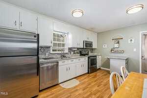 A modern kitchen with wooden flooring, featuring white cabinets, stainless steel appliances, a window above the sink, and a small dining area with a wooden table and chairs. The walls are a light green color with decorative accents.