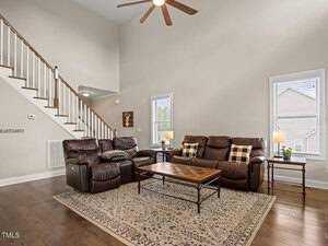 A cozy living room with brown leather sofas, plaid pillows, and a wooden coffee table on a patterned rug. There's a staircase with wooden railings, two windows, and a ceiling fan above. Warm lighting and decor create a welcoming atmosphere.