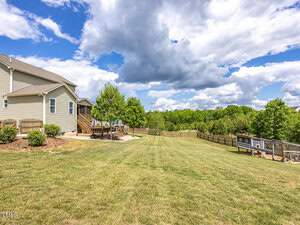 A spacious backyard with a neatly mowed lawn, a wooden fence, and a chicken coop. A house with a deck and trees lines one side, while a partly cloudy sky stretches overhead.