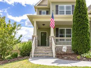 A two-story house with a green front door, stone-accented porch, and large American flag. The porch has white railings and two square columns. A tree and bushes adorn the landscaped front yard under a partly cloudy sky.