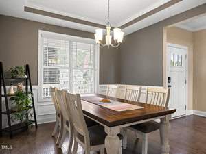 A dining room with a wooden table and eight chairs on a hardwood floor. A hanging chandelier provides overhead lighting. A large window with blinds is on the left wall, and a small black shelf with plants is in the corner. A door is on the right.