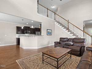 Spacious living room with brown leather sofas, a wooden coffee table, and a patterned rug. Open kitchen with dark wood cabinets and bar seating. High ceiling and a staircase leading to an upper level. Neutral wall colors and hardwood flooring.