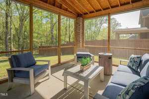 A screened-in patio with wooden ceiling and furnished with two blue cushioned chairs, a loveseat, and a coffee table with a plant. The patio overlooks a fenced backyard with trees visible in the background.