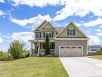 Nestled on 3534 Lavender Lane in Wake Forest, this two-story house boasts green siding and stone accents. A charming front porch with columns and a proud American flag complements the double garage. The home is perfectly situated on a well-maintained lawn under a bright blue sky with scattered clouds.