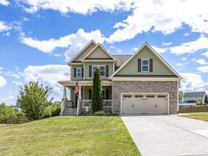 Nestled on 3534 Lavender Lane in Wake Forest, this two-story house boasts green siding and stone accents. A charming front porch with columns and a proud American flag complements the double garage. The home is perfectly situated on a well-maintained lawn under a bright blue sky with scattered clouds.