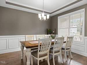 A dining room with a wooden table and eight matching chairs. The walls have white wainscoting and are painted a taupe color. A window with blinds lets in natural light, and a chandelier hangs above the table.