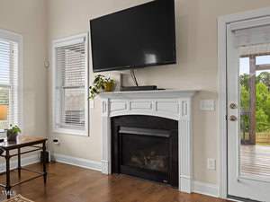 A living room with a wall-mounted TV above a white fireplace. A small plant sits on the mantel. There are two windows and a glass door leading to a deck. The hardwood floor complements the light beige walls.