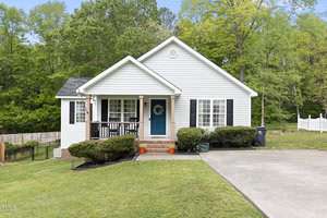 A single-story white house with blue shutters and a teal door, nestled among lush green trees. A small front porch features a decorative wreath. The lawn is neatly mowed, and a paved driveway is visible to the right.