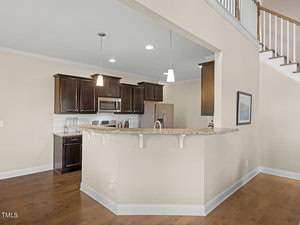 Modern kitchen with dark wood cabinets, stainless steel appliances, and a curved granite countertop. Two pendant lights hang above the counter, and a staircase is visible in the background with a picture on the beige wall.