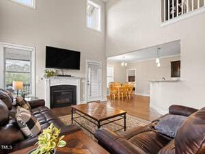 Spacious living room with high ceilings and large windows, featuring leather sofas, a fireplace with a mounted TV, and a wooden coffee table. Adjacent is a dining area with a wooden table, lit by pendant lights. The decor is modern and inviting.