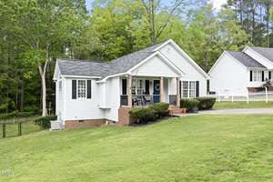 A white, single-story house with a black shingled roof surrounded by green grass and trees. The house has a porch with steps, railings, and bushes in front. A neighboring house and a fence are visible in the background.